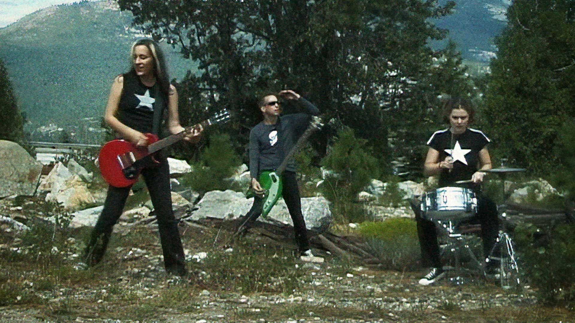 A group of people with guitars and drums performing in front of a mountain at the Groovie Ghoulies concert.