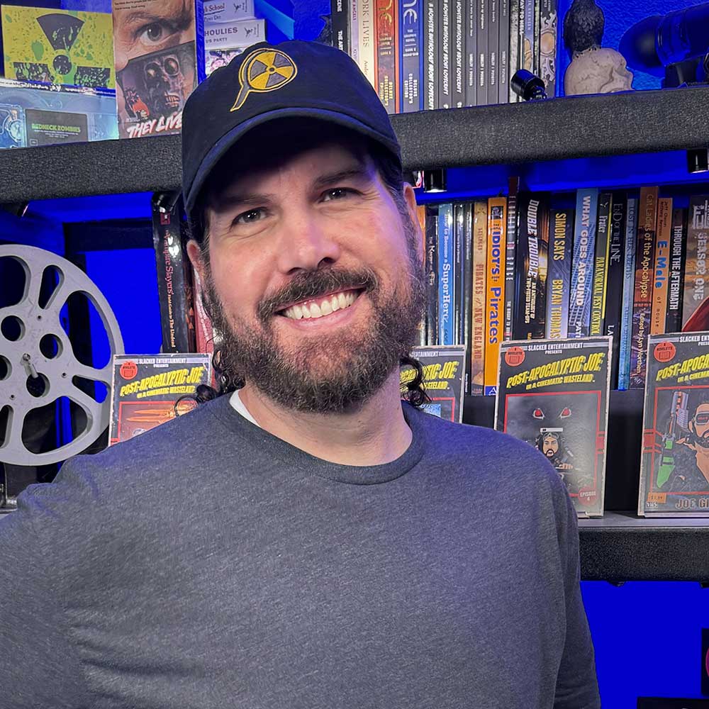 A bearded man in a black hat and gray shirt smiles in front of shelves filled with movies and film reels.